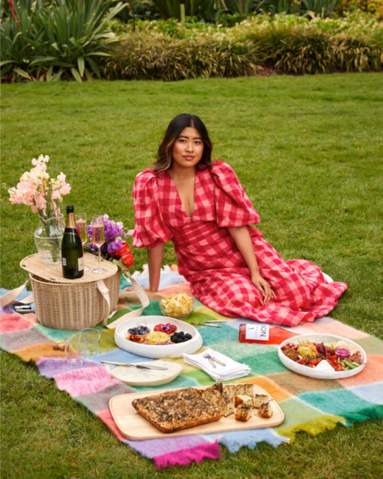 Jessica Nguyen wears a pink and red dress and sits on a picnic blanket surrounded by food and drinks.