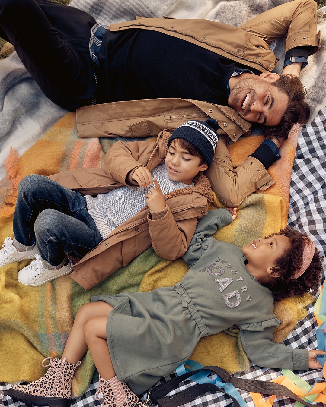 Two children and their dad laying on a picnic blanket, wearing Country Road
